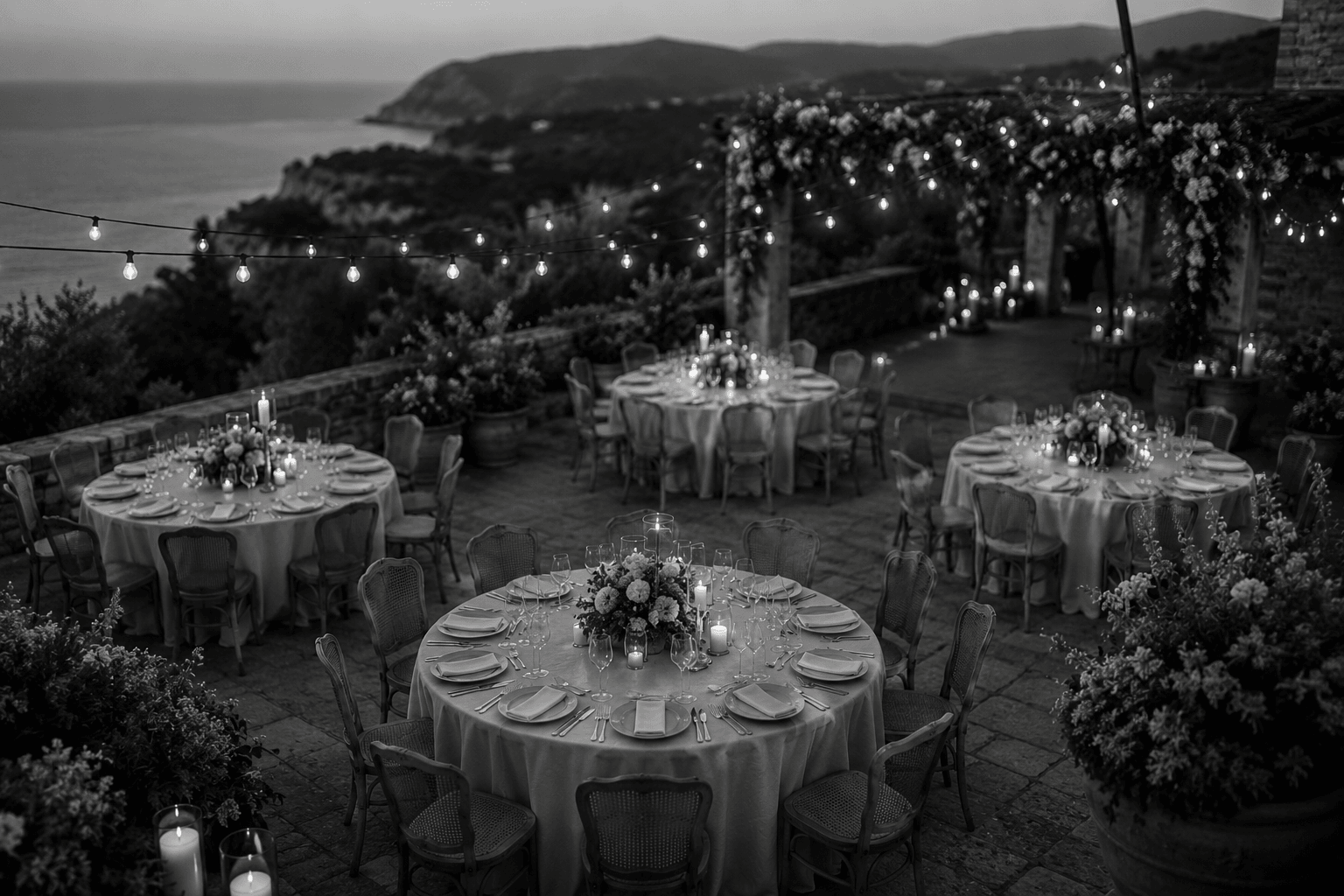Gedeckte Tische mit Kerzen und Lichterketten auf einer Terrasse am Abend, Blick auf die Küste im Hintergrund