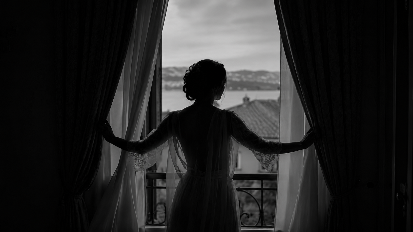 Braut öffnet als Silhouette die Vorhänge vor einem Fenster mit Seeblick
